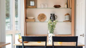 A dining area with a wooden table and chairs, a glass shelf with kitchenware, and a window providing natural light. - The Chester-2 bed Static Caravan (Jedburgh)