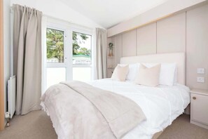 A bedroom with a large bed, a bedside table, a nightstand, a window with curtains, and a view of greenery outside. - The Chester-2 bed Static Caravan (Jedburgh)
