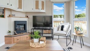 A modern living room with a TV, wooden cabinets, a sofa, and a view of the outdoors. - The Chester-2 bed Static Caravan (Jedburgh)