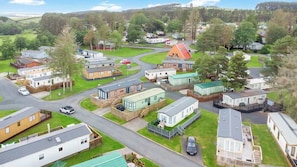 A residential area with mobile homes, a main road, and surrounding greenery. - The Chester-2 bed Static Caravan (Jedburgh)