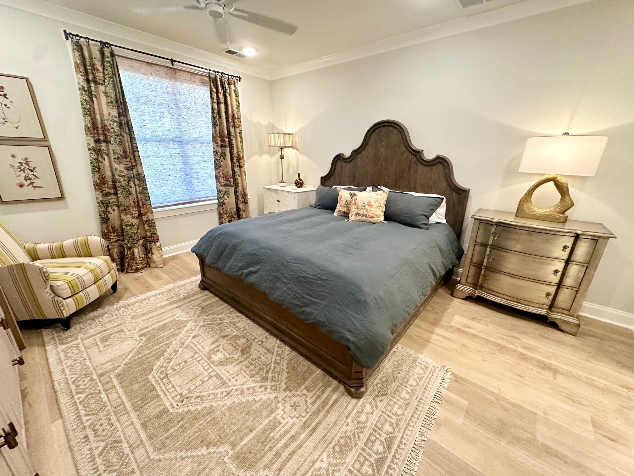 A bedroom with a large bed, a chair, a nightstand, a dresser, and a window with curtains.