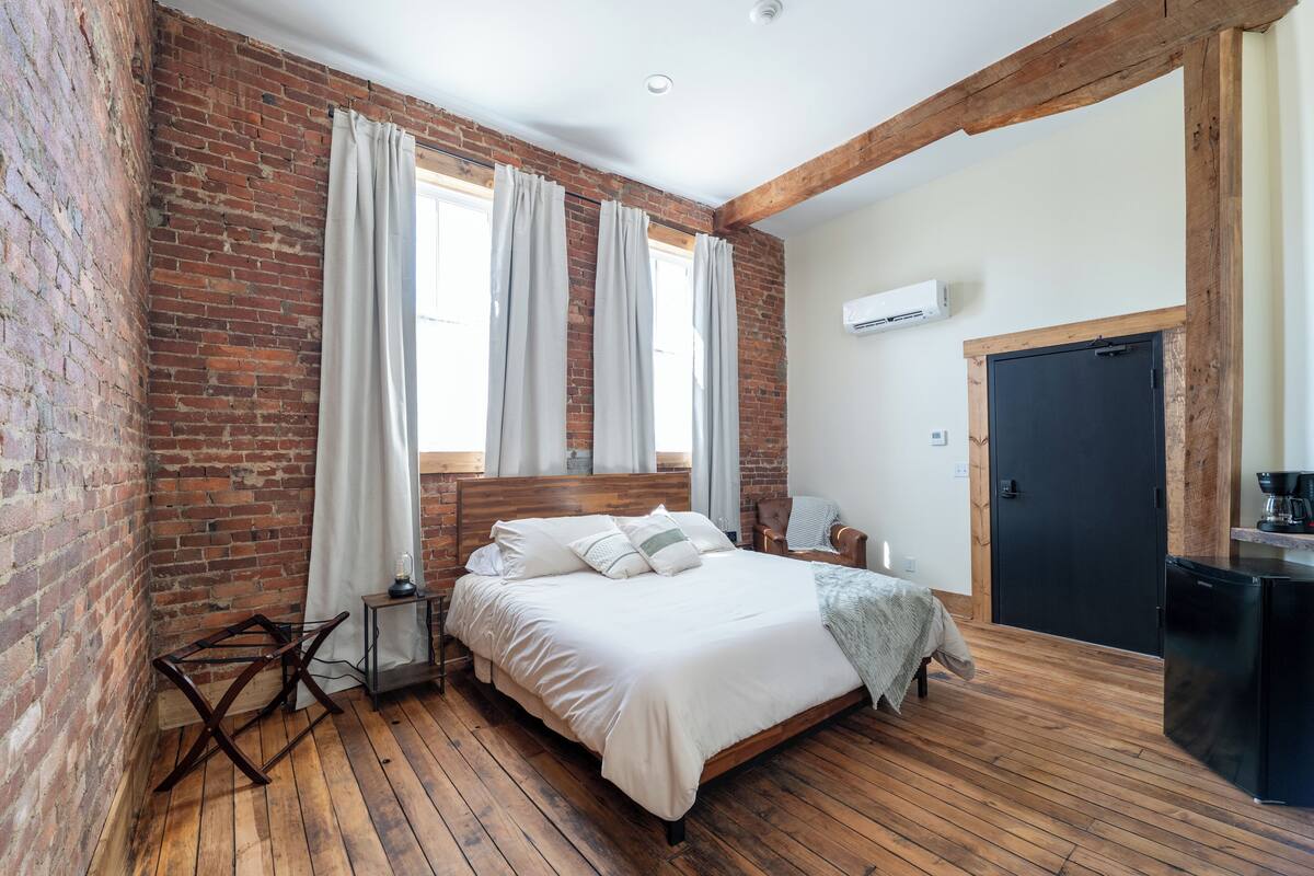 A bedroom with a brick wall, wooden floor, a bed with white bedding, a small bedside table, and a black wardrobe.