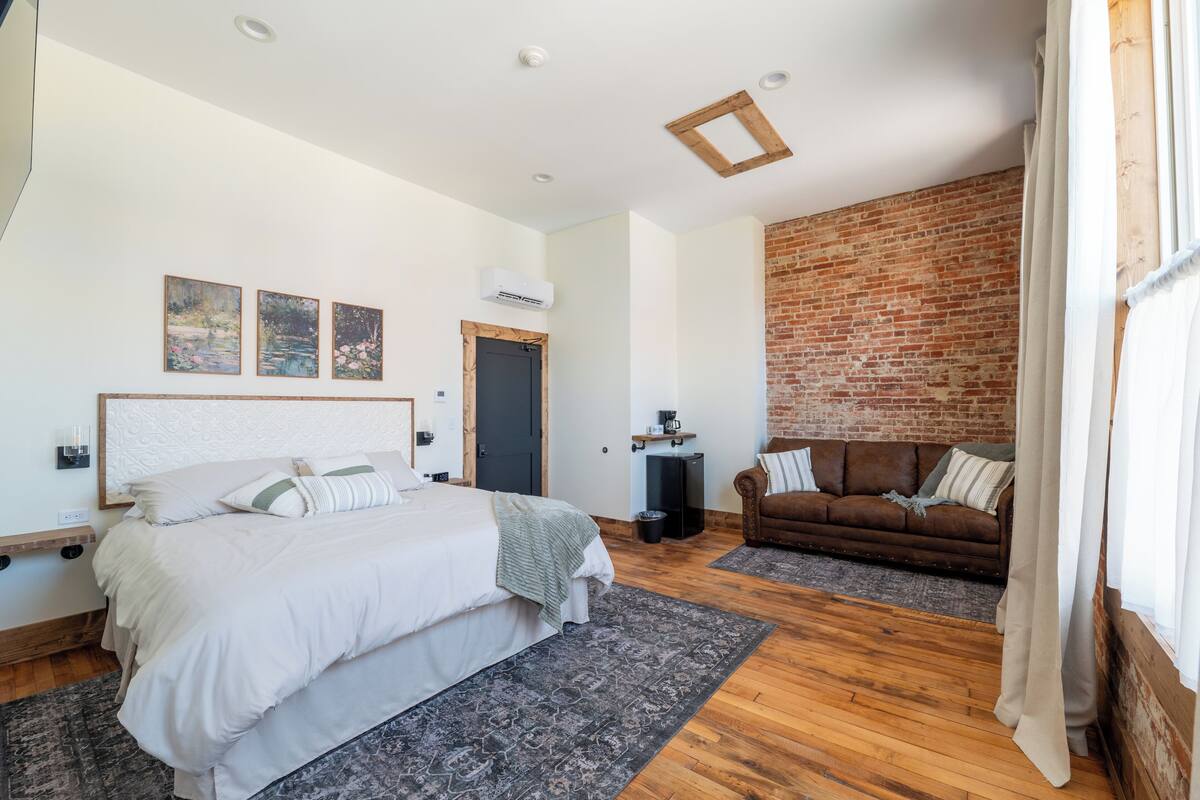 A bedroom with a large bed, a brown leather sofa, a brick wall, and a ceiling with recessed lighting.