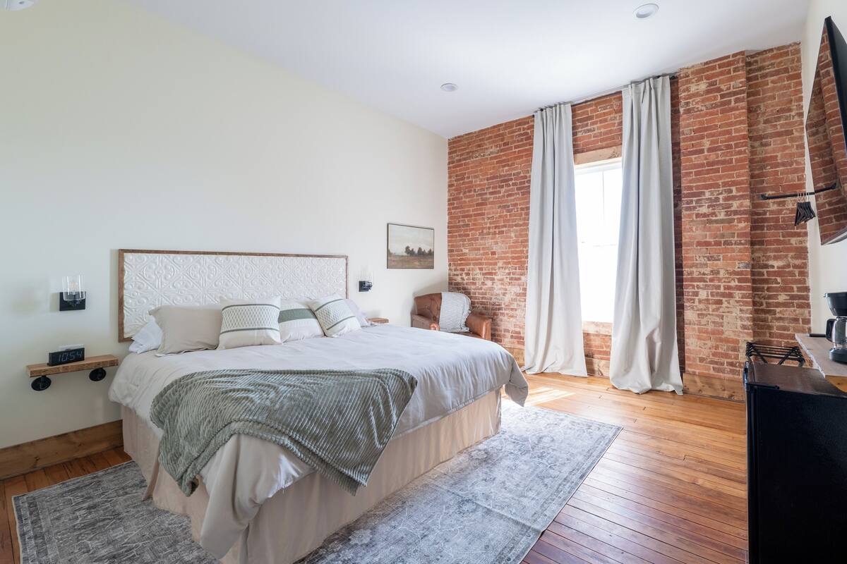 A bedroom with a large bed, a brick wall, a window with curtains, a flat-screen TV, and a nightstand with a lamp.