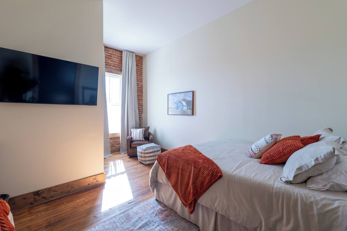 A bedroom with a large bed, a TV mounted on the wall, and a window with curtains.