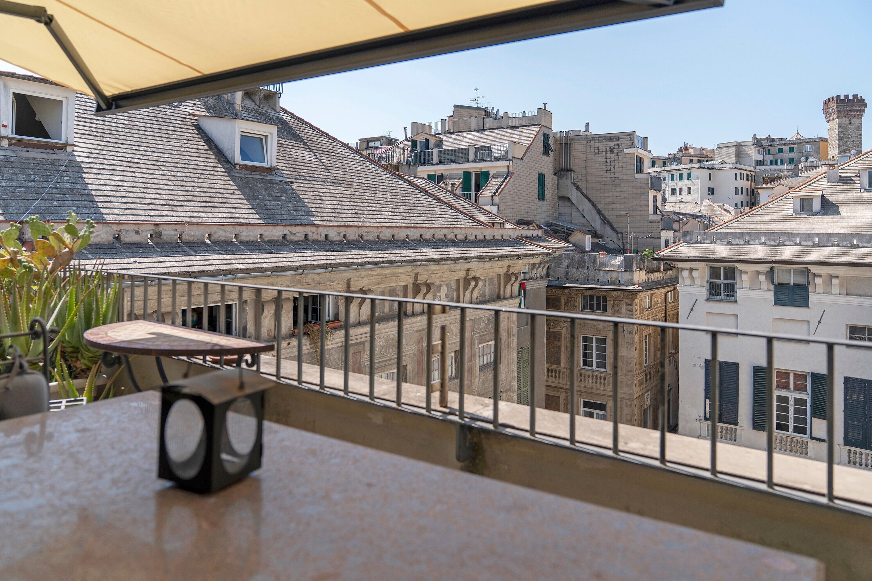 A balcony with a table, chairs, and a speaker, overlooking a cityscape with historic buildings and a tower.