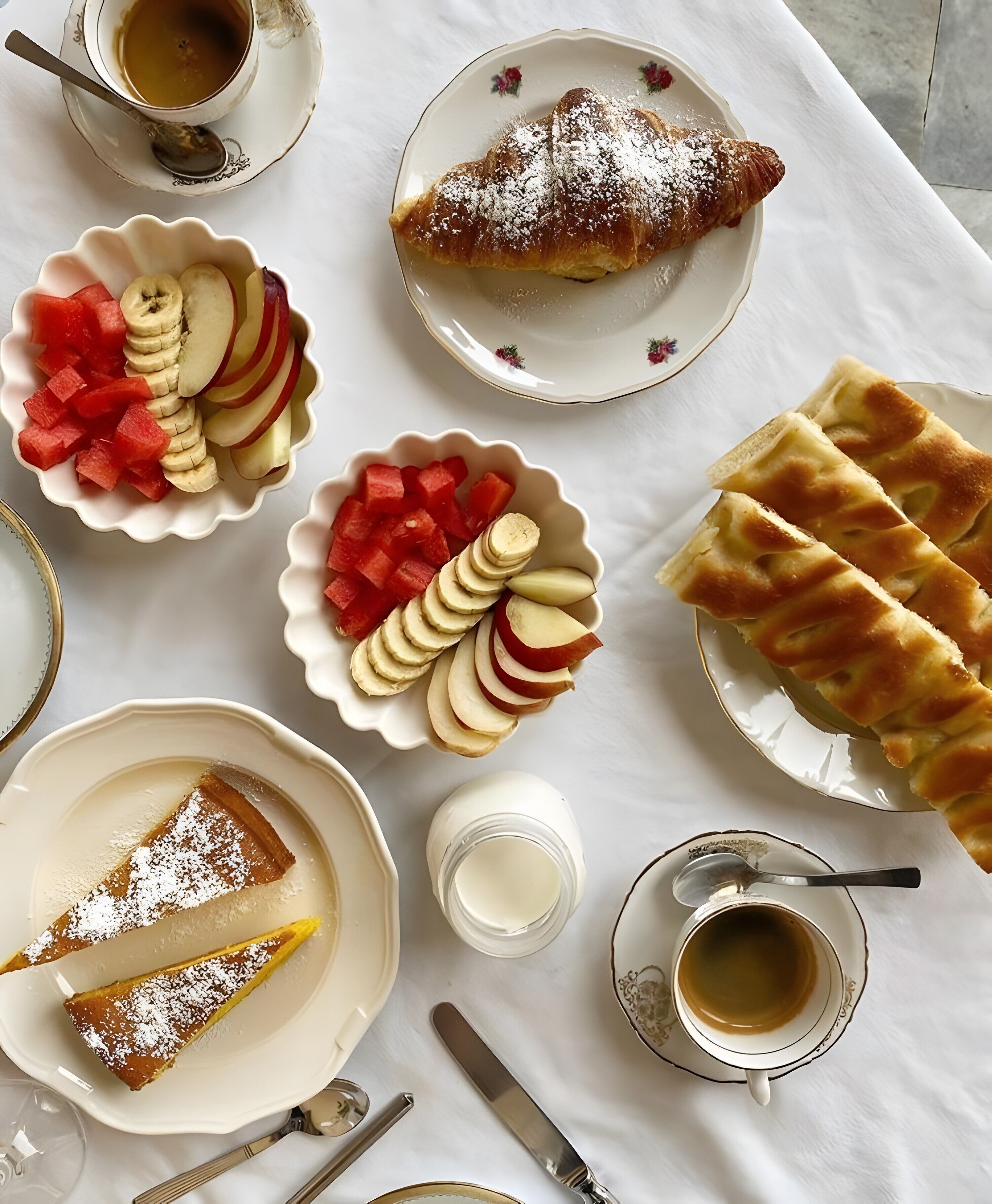 Une table sur laquelle on trouve divers aliments et boissons, notamment un croissant, des tranches de pomme, de la pastèque et une part de tarte.