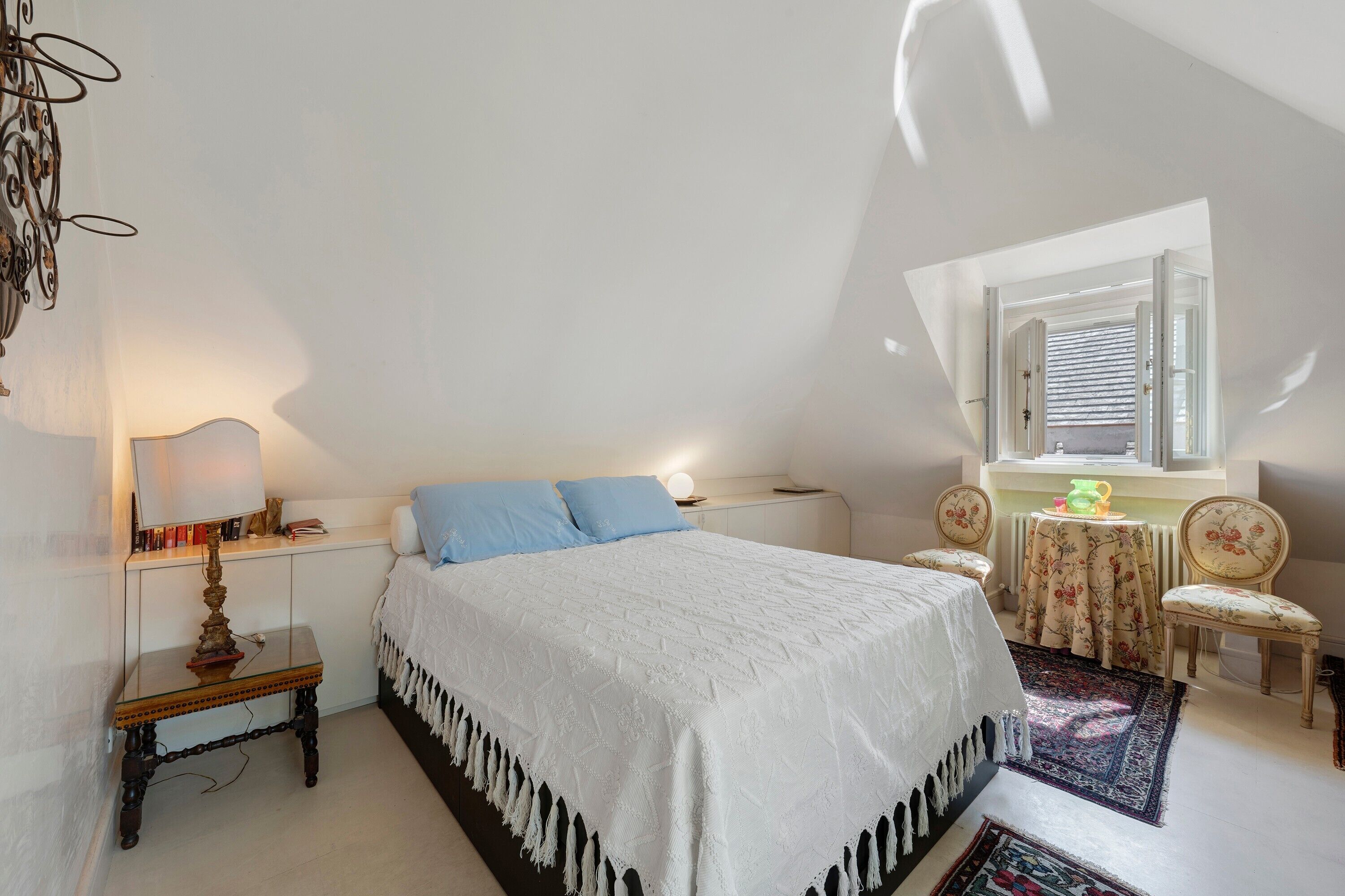 Une chambre à coucher comprenant un lit, une table de chevet, une lampe, une chaise et une fenêtre avec des stores.