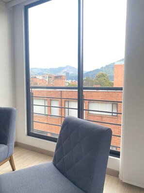 Une pièce dotée d’une grande fenêtre, d’un fauteuil bleu et offrant une vue sur des immeubles et des montagnes.