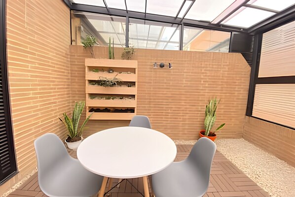 Un espacio al aire libre moderno con una mesa blanca y dos sillas, rodeado de muros de ladrillo y plantas en macetas.