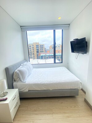 Une chambre avec une grande fenêtre, un lit, un téléviseur fixé au mur et une table de chevet avec une lampe et une télécommande.