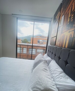 A hotel room with a large bed, a view of buildings and mountains through a window, and a modern headboard. - Edificio Benyalt 94 (Bogota)