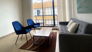 A modern living room with a grey sofa, two blue chairs, a wooden coffee table, and a large window with a view of a building. - Edificio Benyalt 94 (Bogota)