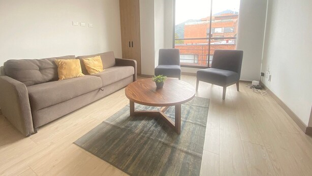 A modern living room with a sofa, two armchairs, a coffee table, and a large window with a view of buildings. - Edificio Benyalt 94 (Bogota)