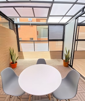 Un patio interior moderno con una mesa redonda y dos sillas, plantas en macetas y vistas amplias a través de grandes ventanales.