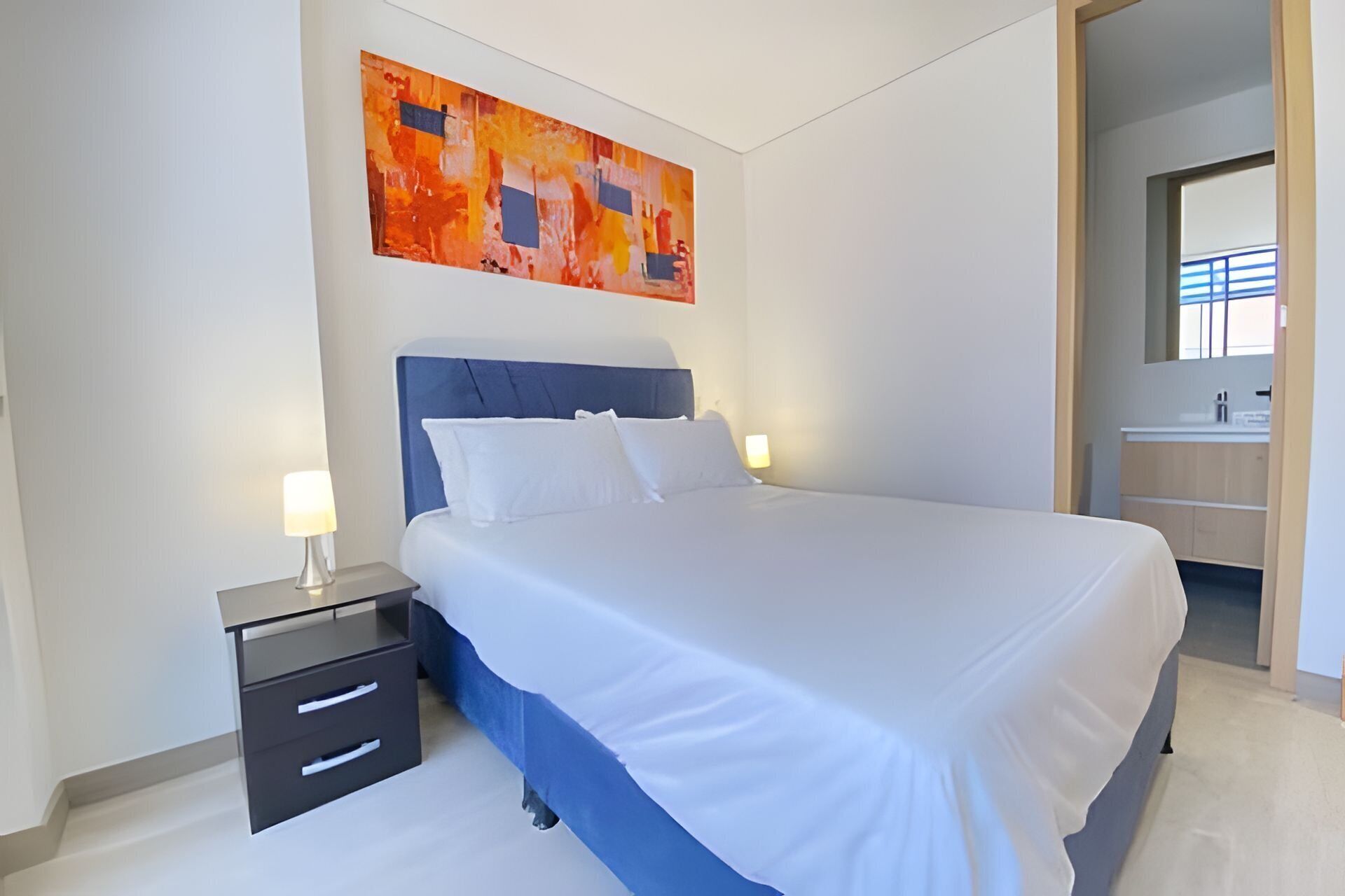 Un dormitorio con cama, mesitas de noche, un cuadro colgado en la pared y un baño visible a través de un puerta abierta.