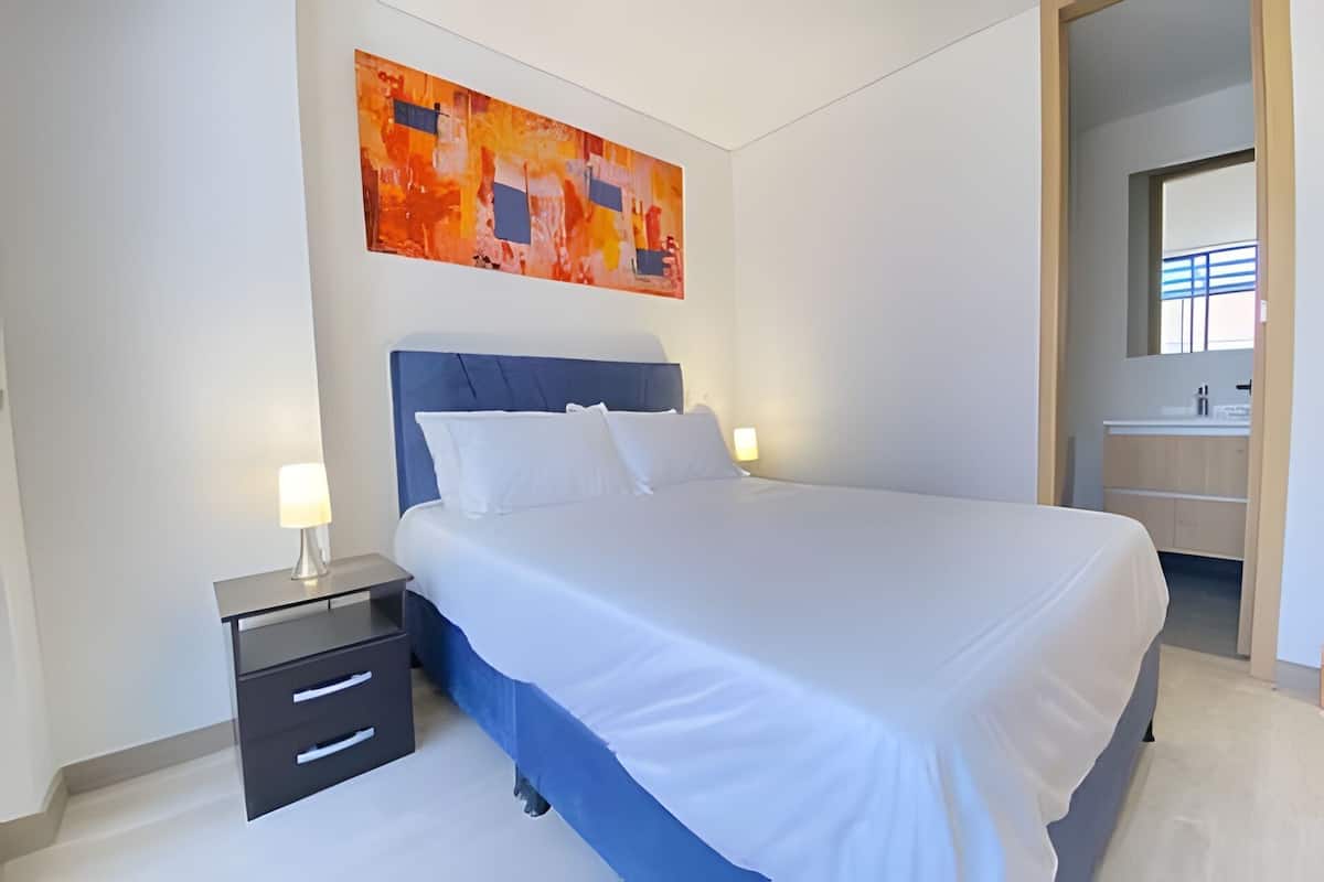 Un dormitorio con cama, mesitas de noche, un cuadro colgado en la pared y un baño visible a través de un puerta abierta.