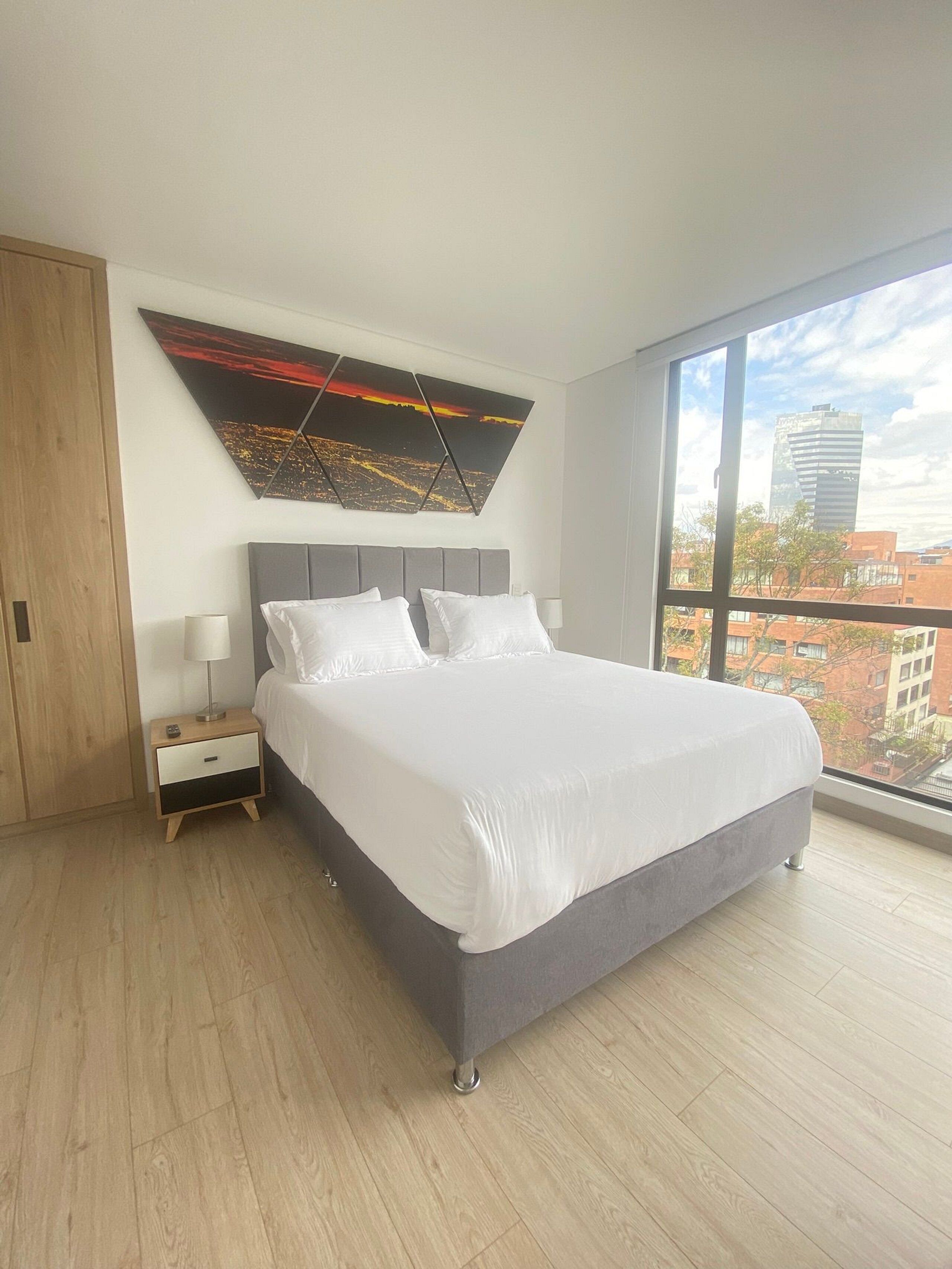 Un dormitorio moderno con una cama grande, una mesita de noche, una obra de arte en la pared y un ventanal con vistas a la ciudad.