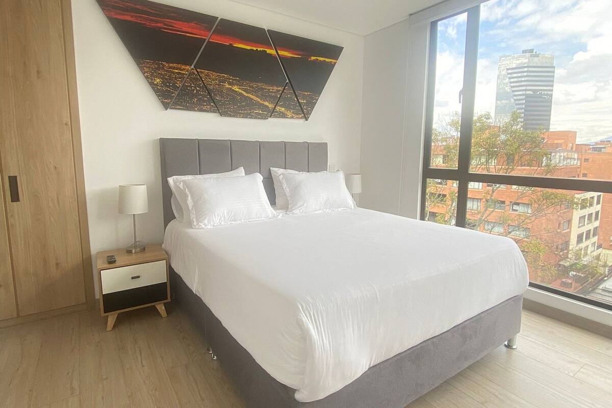 Un dormitorio moderno con una cama grande, una mesita de noche, una obra de arte en la pared y un ventanal con vistas a la ciudad.