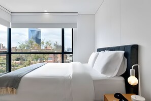 Habitación de hotel moderna con un ventanal grande, una cama, una mesita de noche con lámpara y vistas a la ciudad.