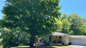 A single-story house with a porch, a garage, and a large tree in front. - Charming 2BR near Lake Barkley – fully equipped and ready for your stay! (Grand Rivers)
