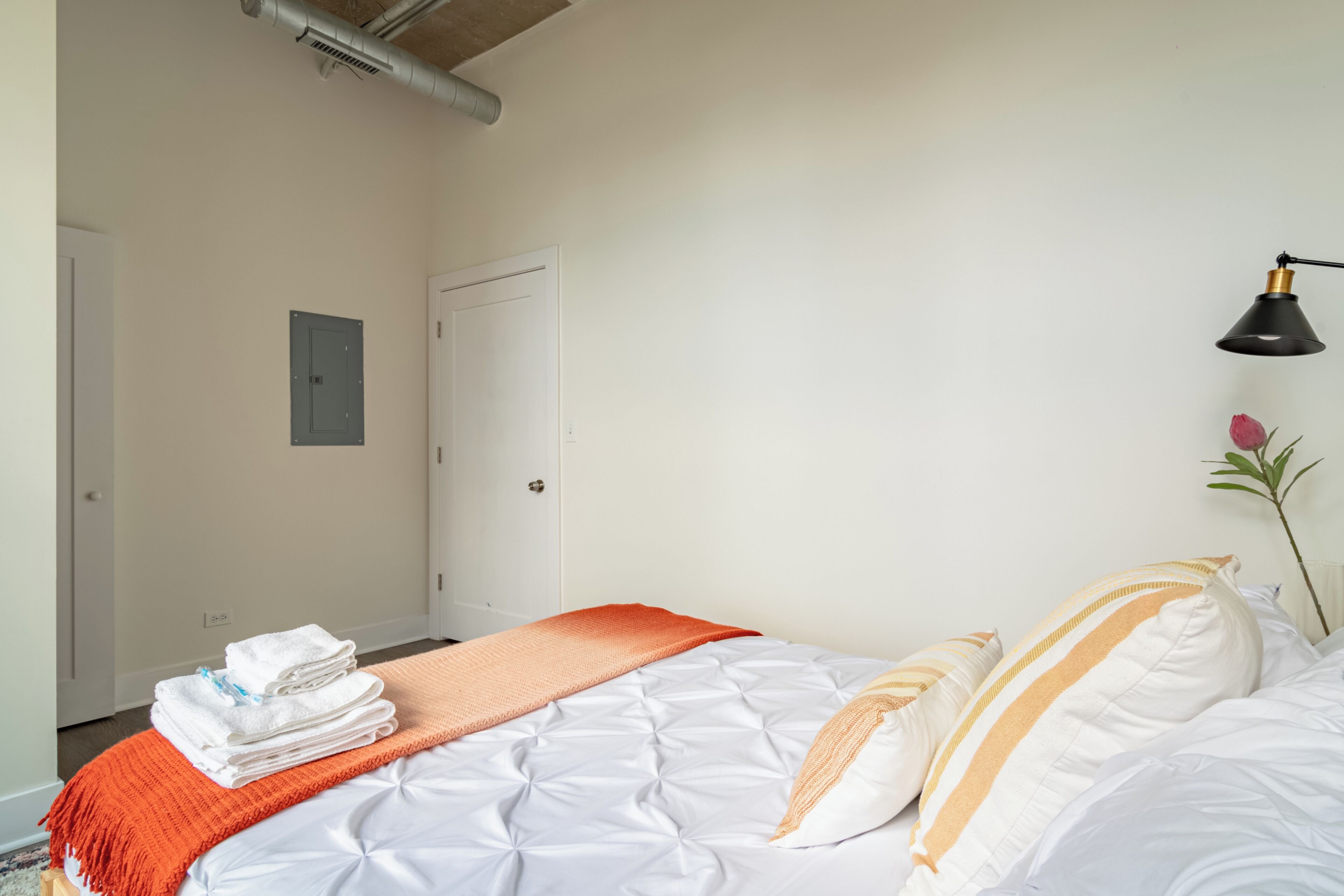 Ein ordentlich bezogenes Bett mit weißen Bettwäsche und einer orangefarbenen Decke, eine schwarze Schreibtischlampe und ein Blumentopf auf der rechten Seite.