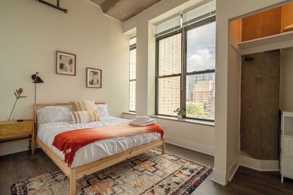 A bedroom with a large bed, a window with a city view, and a wooden floor. - Skyline 2BR in South Loop Chicago Near McCormick Place & Soldier Field (Chicago)