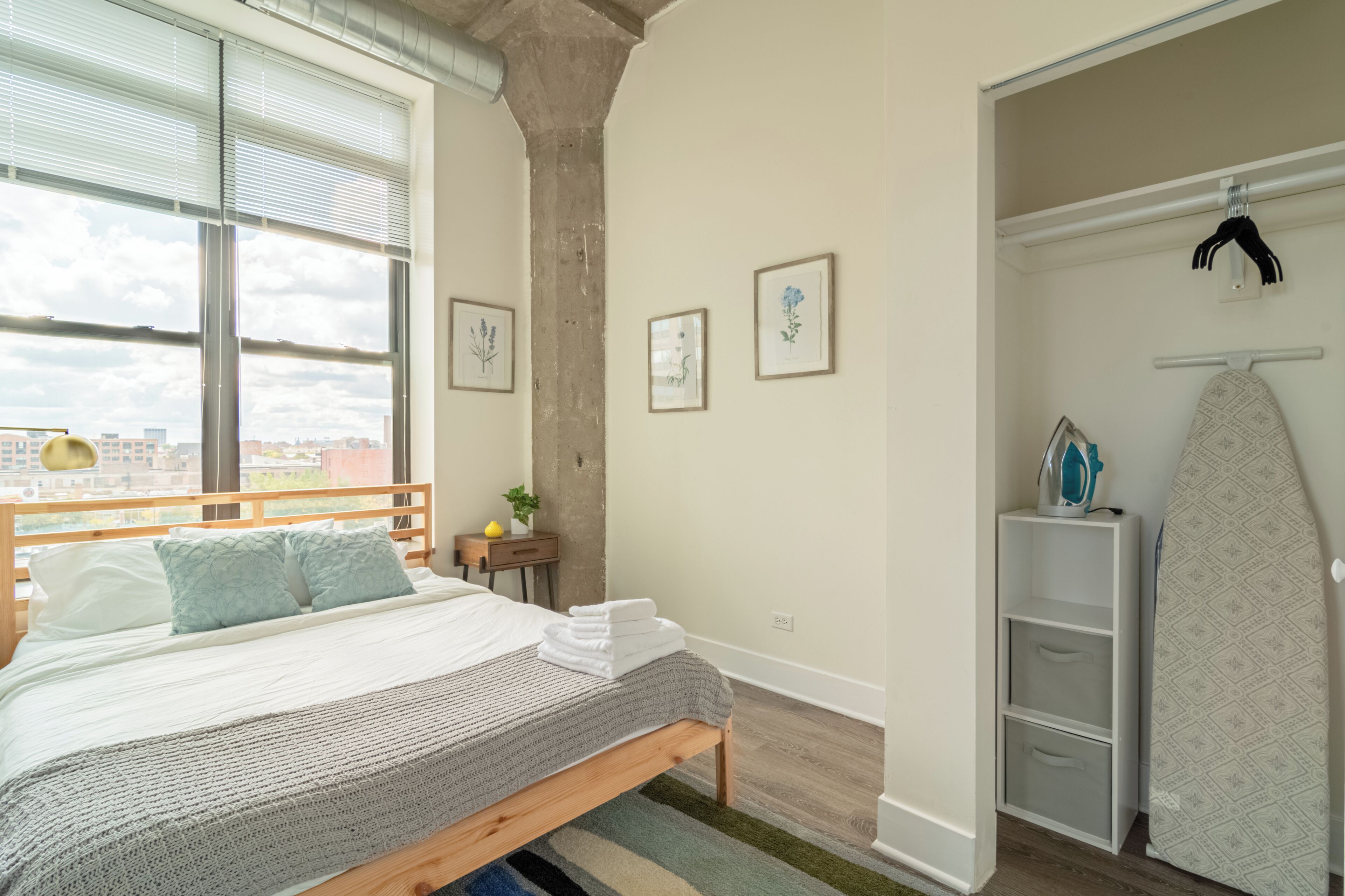 A bedroom with a bed, a wooden headboard, a window with blinds, a small table with a plant, and a wall-mounted ironing board.