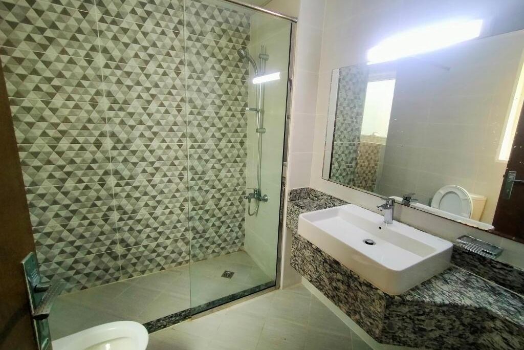 Un baño moderno con pared de ducha revestida de azulejos geométricos, ducha con mampara de vidrio, lavamanos blanco con encimera de granito y un espejo grande.
