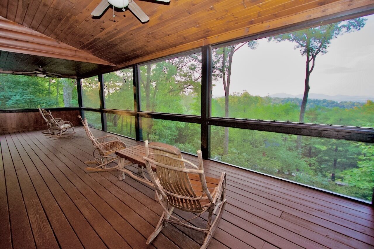 A wooden deck with rocking chairs and a table, large windows offering a view of a forested landscape.