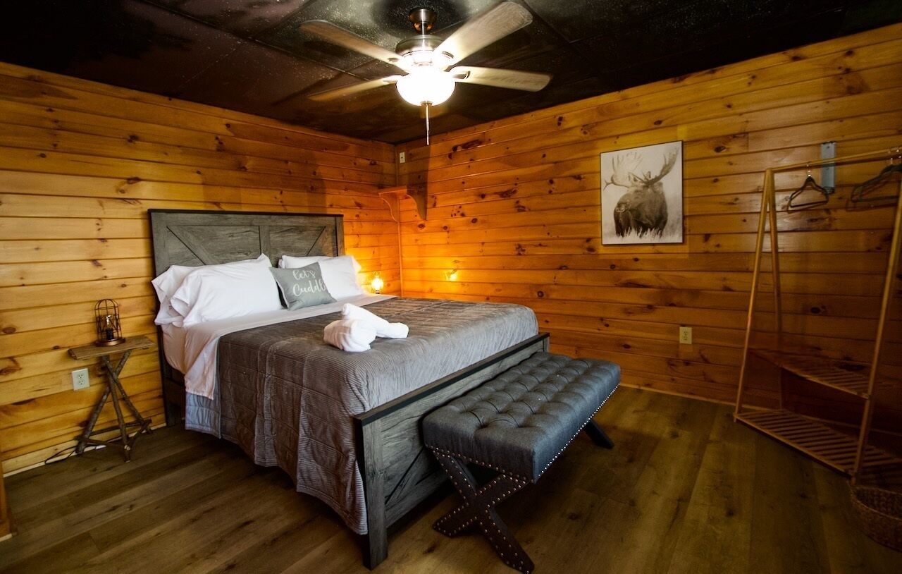 Ein Schlafzimmer einer Holzhütte mit Bett, Sitzbank, Deckenventilator und einem gerahmten Bild von Tieren an der Wand.