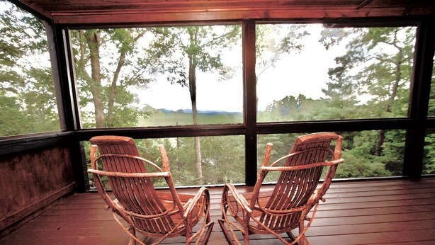 Een houten terras met twee schommelstoelen gericht op een groot raam met uitzicht op een bebost gebied.
