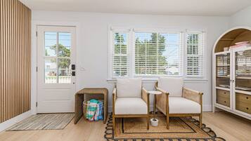 A room with a door, two windows with blinds, a wicker basket, two white chairs, a small table, and a cabinet with glass doors.