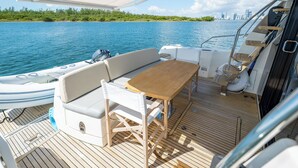 A boat deck with a wooden table and white cushioned seating, a small inflatable boat, and a view of a city skyline across the water. - Amazing 3 bedrooms luxury yacht in North Miami (North Miami)