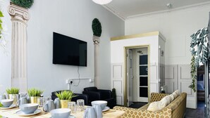 Ein modernes Wohnzimmer mit Essbereich, einem an der Wand montierten Flachbildfernseher und einer Couch mit gemusterten Kissen.