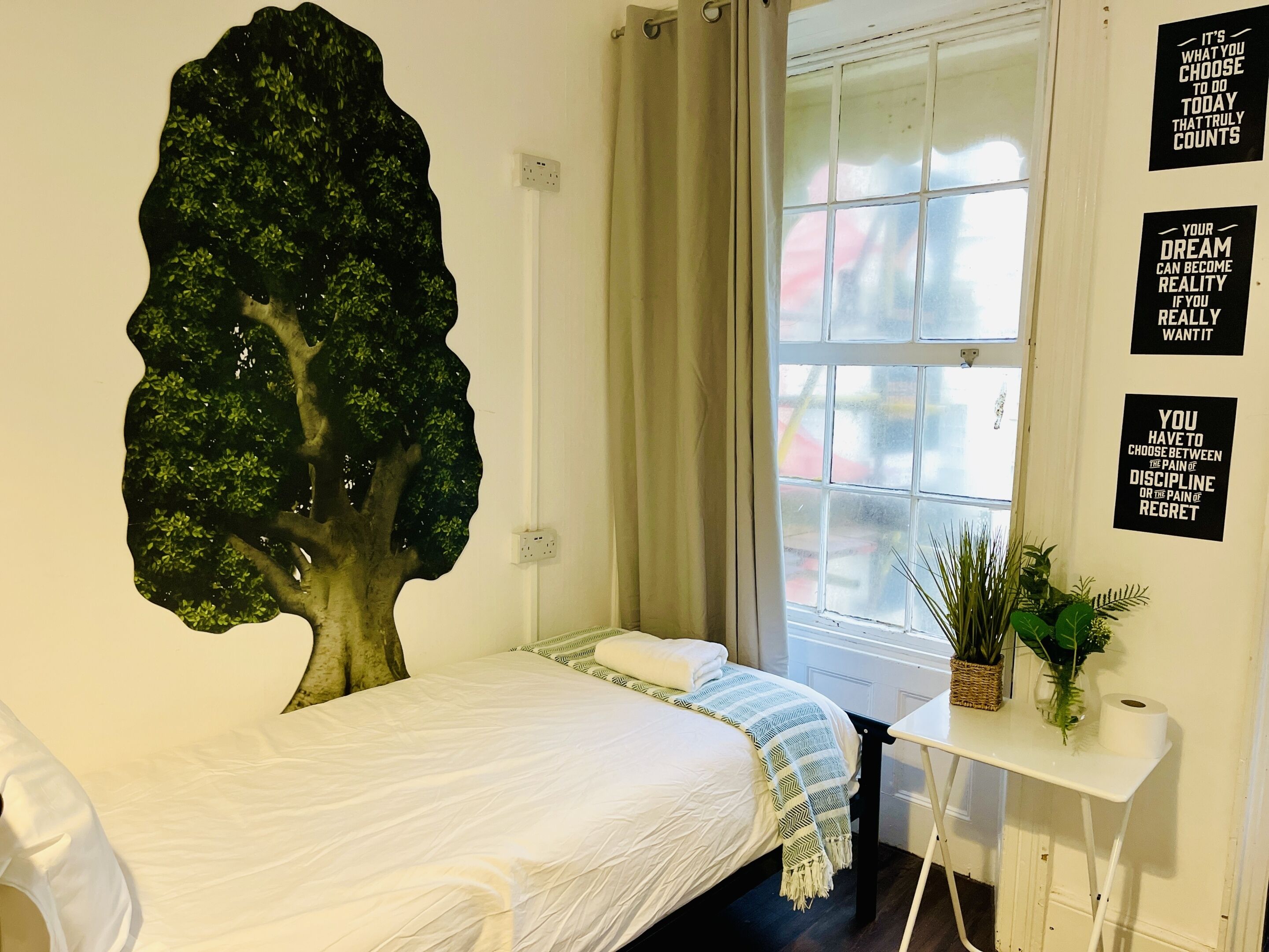 Un dormitorio con una cama, un adorno de árbol en la pared, una ventana con cortinas y carteles motivacionales.