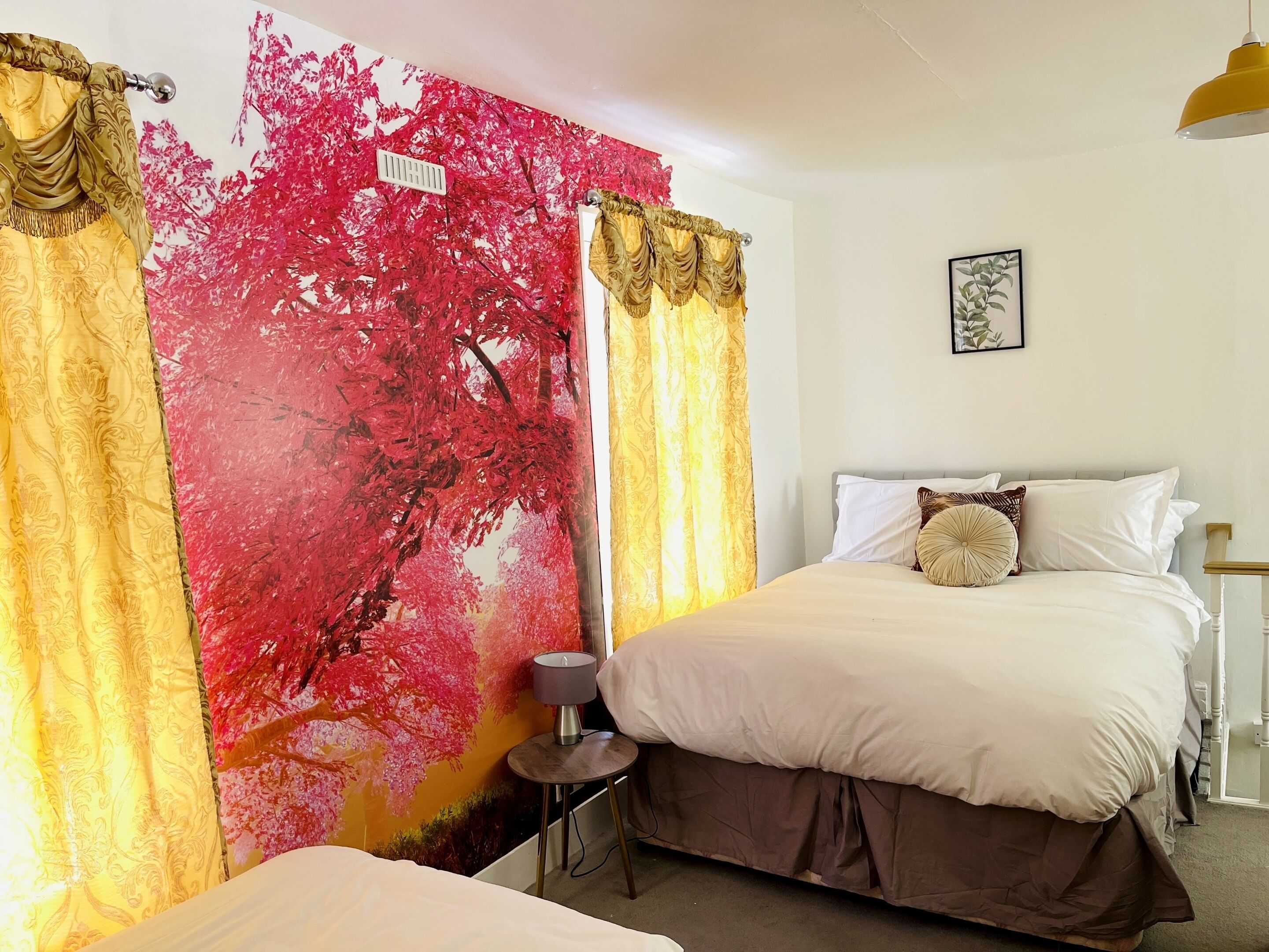 A hotel room with two beds, a red and yellow wall mural, a small table with a lamp, and a framed picture on the wall.