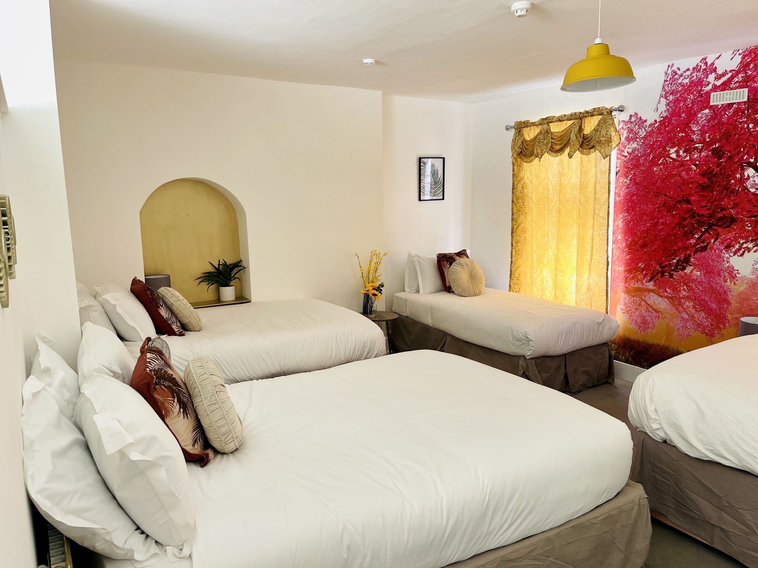 Une chambre d’hôtel avec deux lits simples, une petite table avec une plante, un luminaire suspendu jaune et une décoration murale florale rouge.