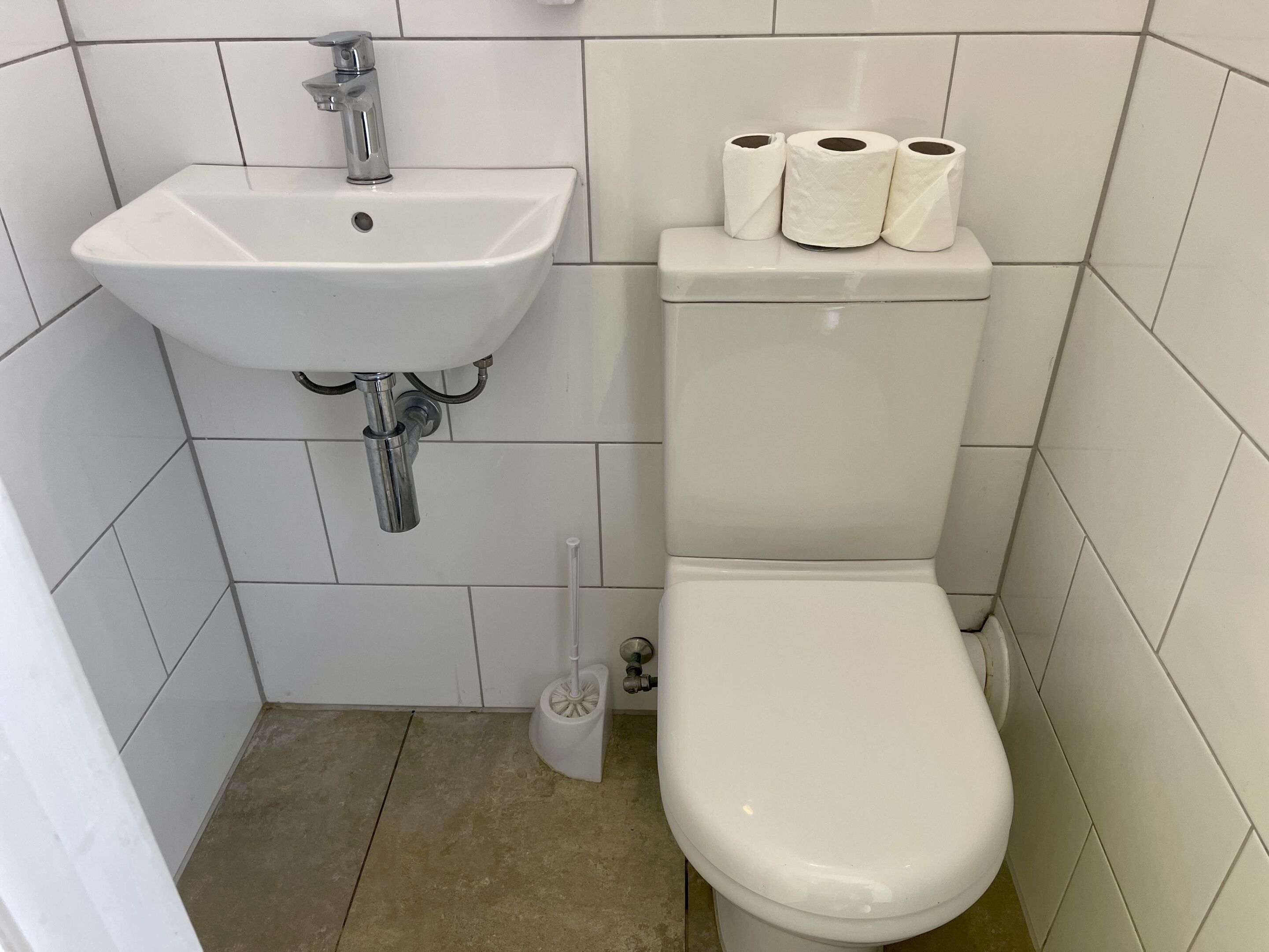 Un baño pequeño con un lavamanos blanco, inodoro y tres rollos de papel higiénico sobre el tanque.