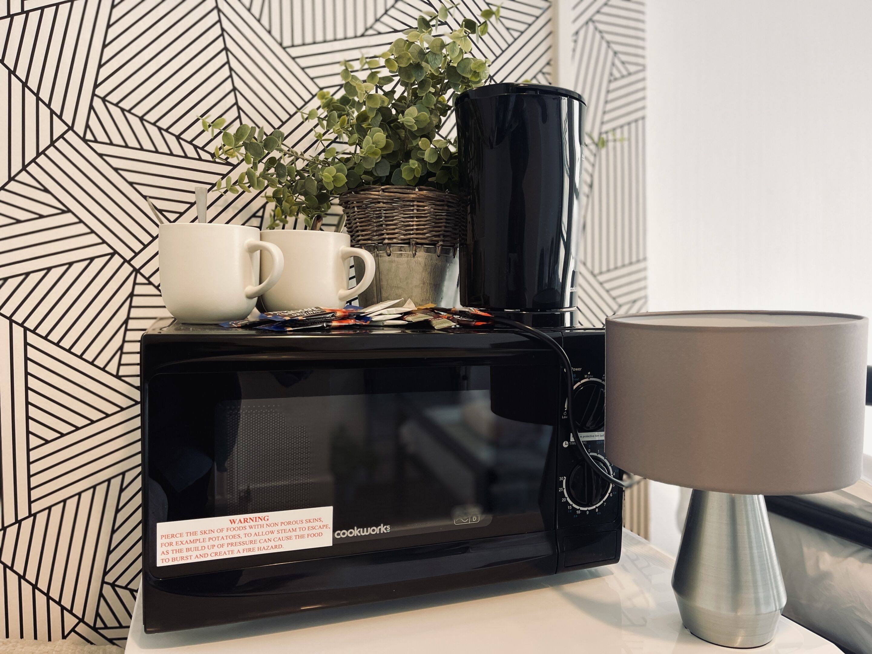 A microwave with a warning label, a lamp, and a plant on a table.