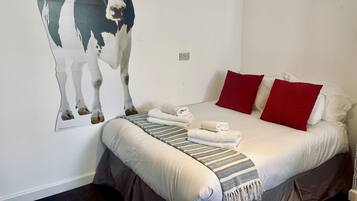 A bedroom with a bed, red and white pillows, a white wall, and a large cow painting.