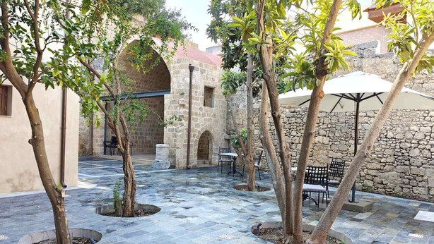 An outdoor courtyard with stone flooring, trees, benches, and a stone wall. - Fabrizio del Carretto (Rhodes)