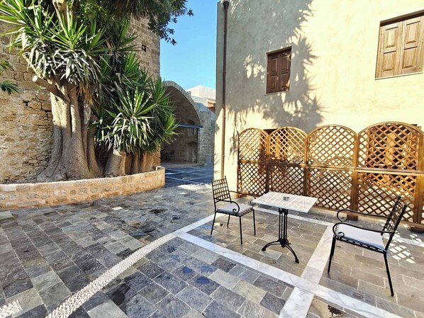A courtyard with stone flooring, a table and chairs, a wooden fence, and a building with a window.