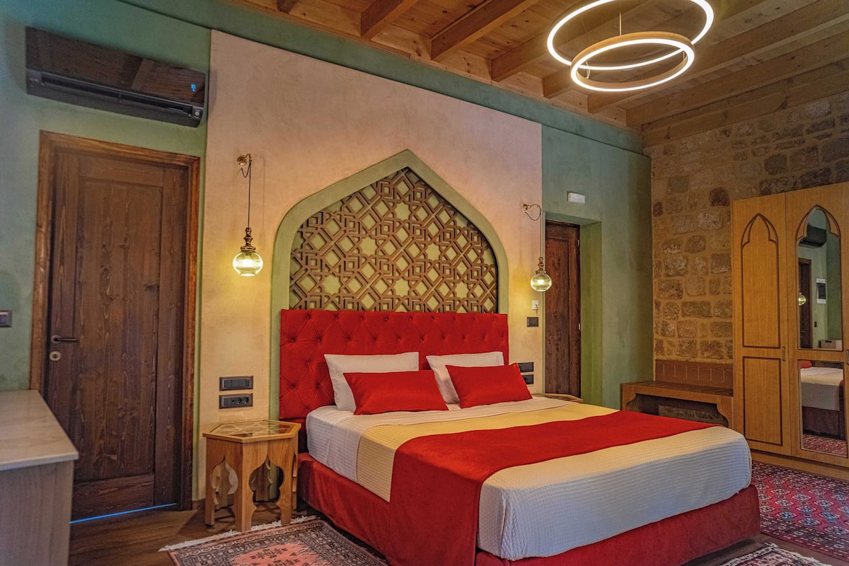 A bedroom with a red bed, wooden headboard, and a stone wall. There is a wooden wardrobe and a nightstand with a lamp.