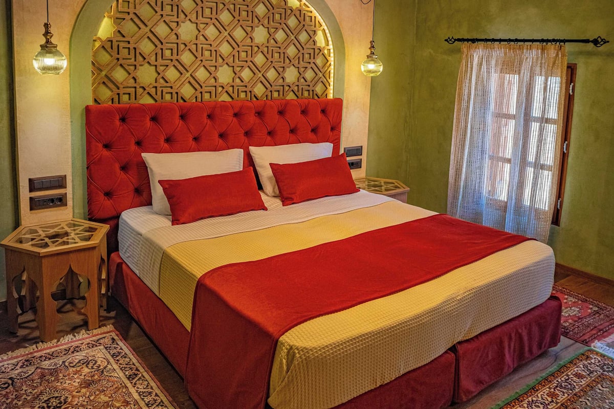 A bedroom with a red tufted headboard, a patterned wall, and a large bed with red and white bedding. There are two bedside tables with lamps, a window with sheer curtains, and a patterned rug on the floor.