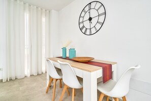 Uma sala de jantar com mesa de madeira, cadeiras brancas, um relógio de parede grande e uma janela com cortinas brancas.