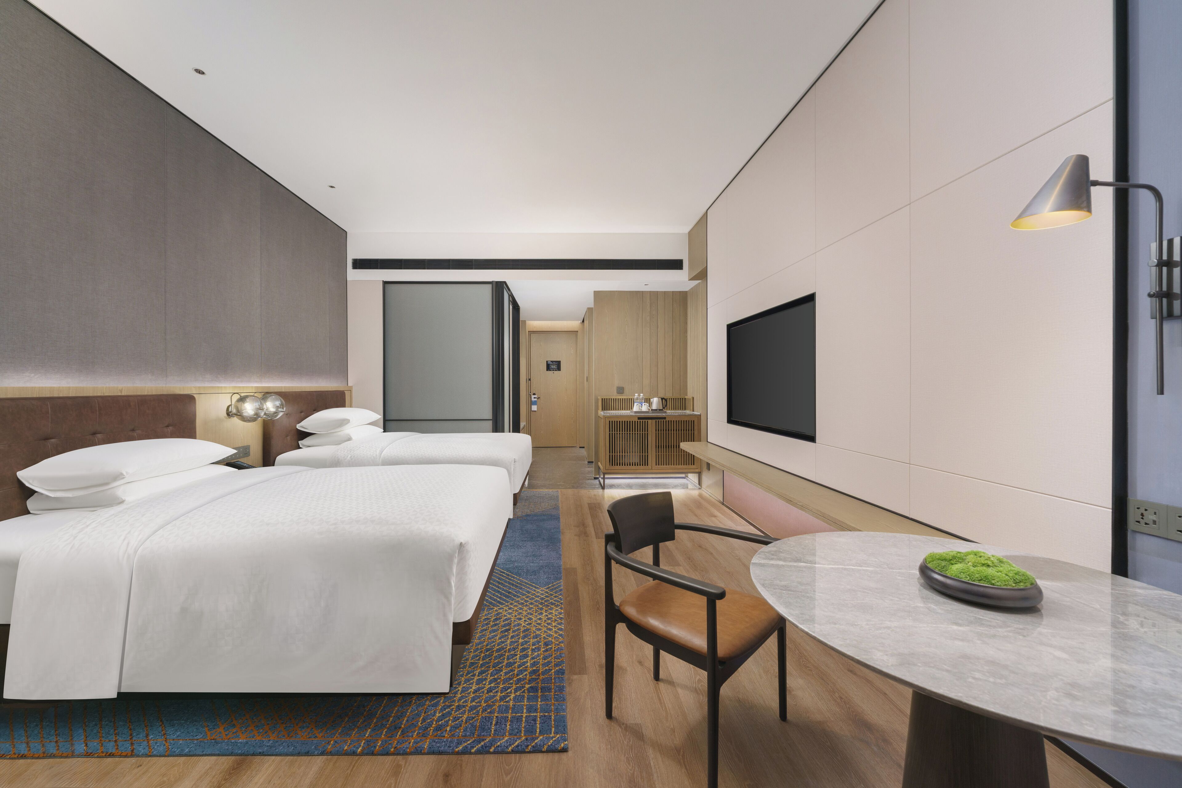 Una habitación de hotel moderna con dos camas, un escritorio, una silla, un televisor y una pequeña mesa con un tazón de sustancia verde.
