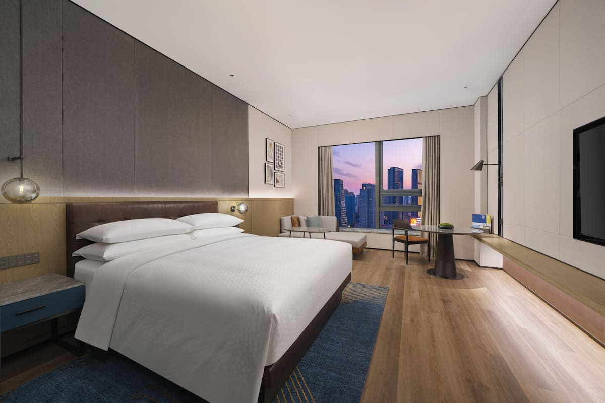 A modern hotel room with a large bed, a desk, and a view of the cityscape through the window.
