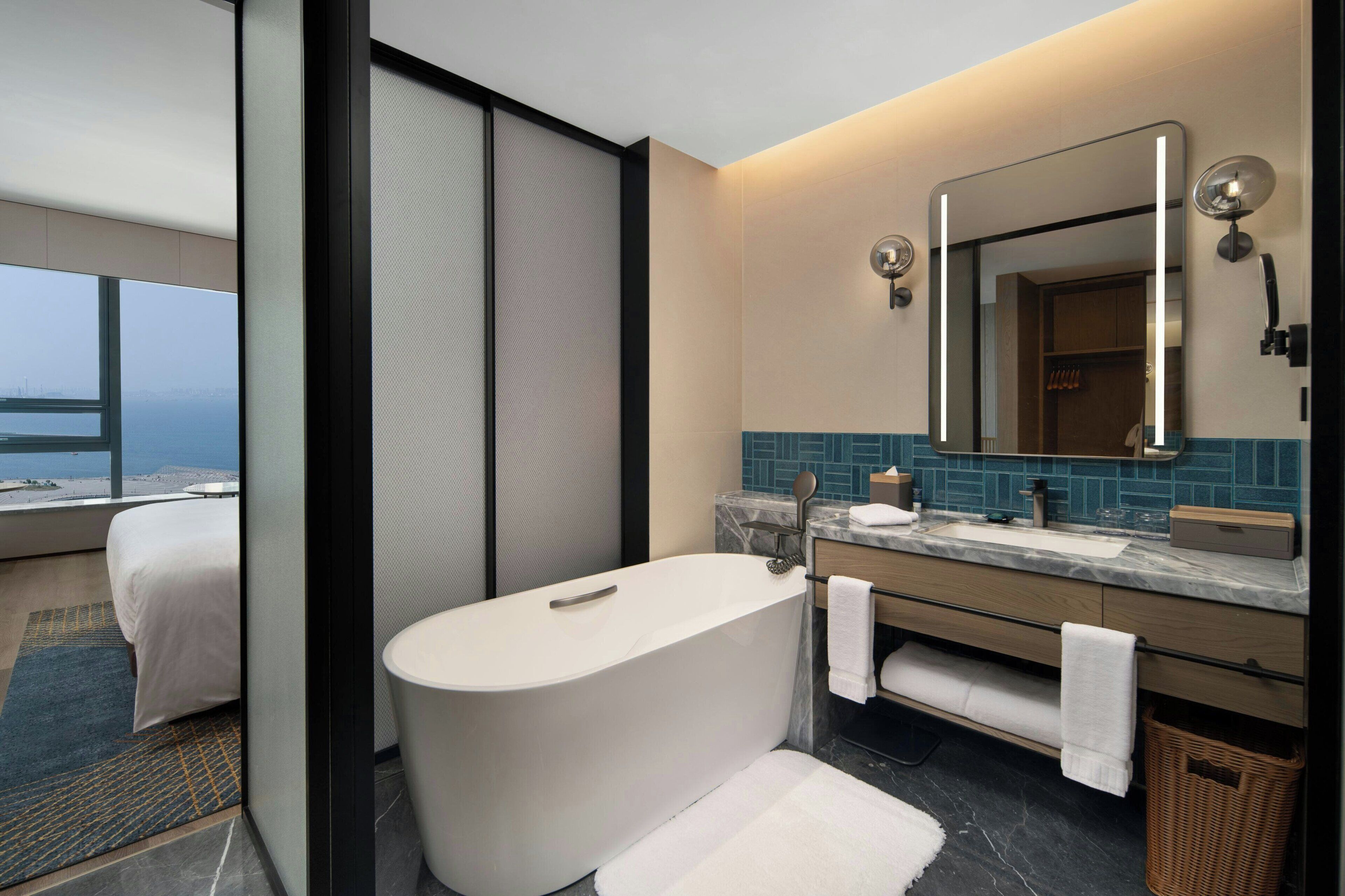 A modern hotel bathroom with a large bathtub, a vanity with a mirror, and a view of the sea through a large window.