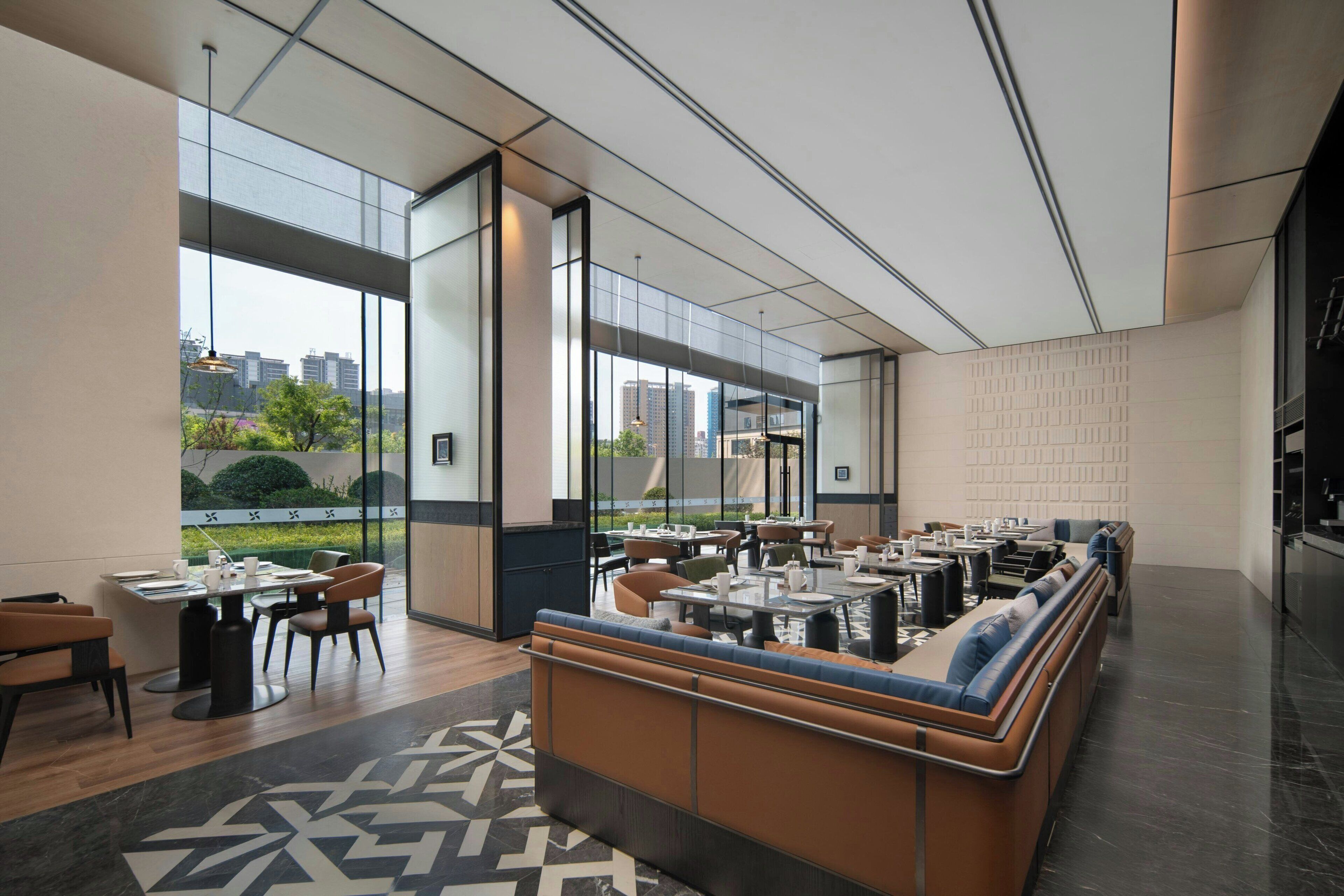 Un espacio interior moderno con una larga zona de asientos tipo banco, mesas y sillas, y grandes ventanales con vistas a la ciudad.